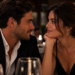 A man in a dark blazer and a woman in a black dress lean toward each other across a candlelit restaurant table, gazing into each other's eyes, with empty wine glasses in the foreground and soft bokeh lighting behind them.