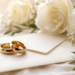 Two gold wedding bands rest on a sealed cream envelope, with white roses, baby's breath, and eucalyptus sprigs arranged on soft ivory fabric behind them.